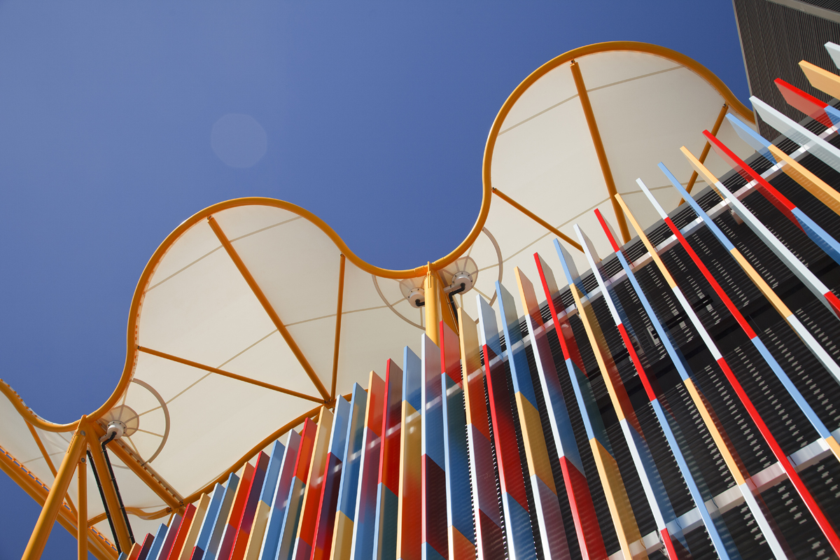 Upward view of People First Stadium roof canopy with colourful vertical façade panels against a clear blue sky.
